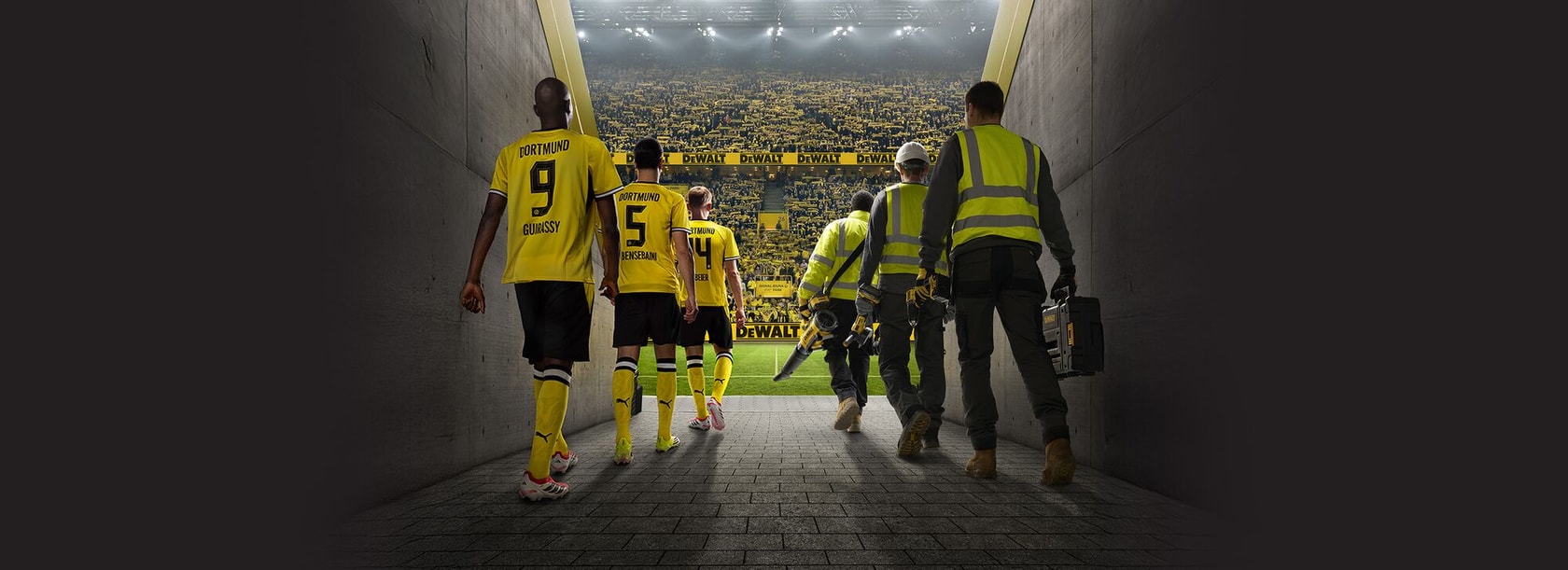 Fußballspieler und Arbeiter in Gelb betreten einen Stadiontunnel zu Fans mit DEWALT-Bannern.
